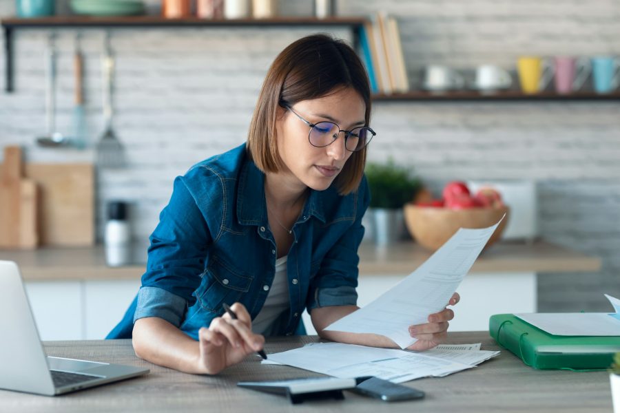 young woman working with computer while consulting some invoices and documents in the kitchen